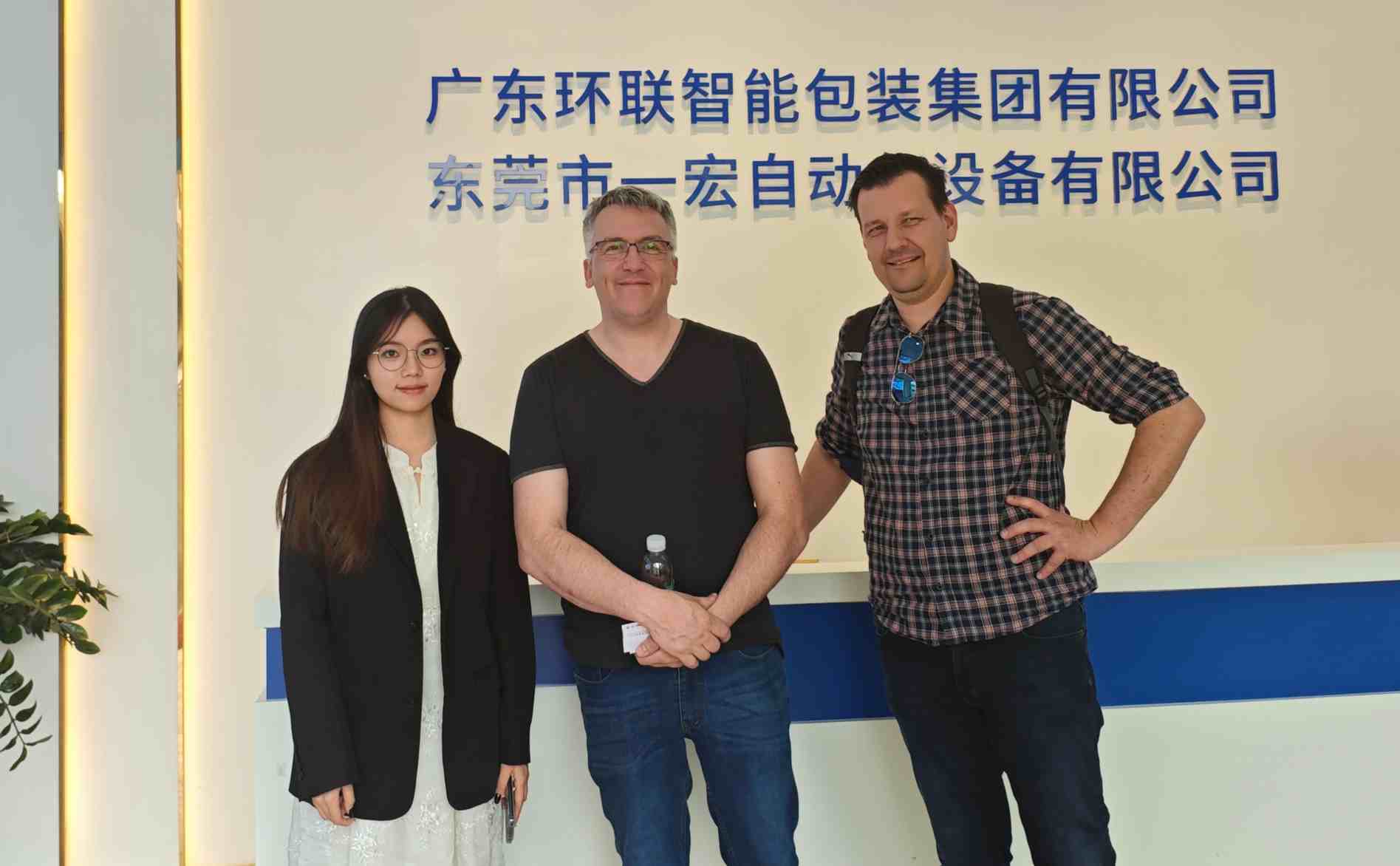 UBL staff member posing for a photo with Australian clients at the front desk area, with the signage of Guangdong UBL Intelligent Packaging Group and Dongguan Yihong Automation Equipment Co., Ltd. displayed in the background