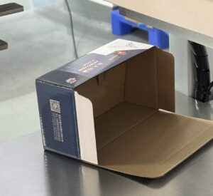 Open e-commerce corrugated packaging box placed on a cartoning machine’s stainless steel platform — used in e-commerce supply chain packaging lines for automatic box forming