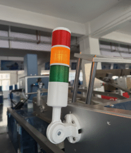 Three-color signal tower light (red, yellow, green) mounted on an automated cartoning machine in a packaging factory, indicating equipment status (operation, warning, shutdown)