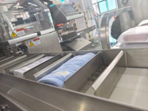 Work station of a fully automatic bagging machine: Folded blue and white towels are placed in the metal trough, ready for bagging. The equipment is fitted with safety warning labels, and the background shows a workshop production environment.