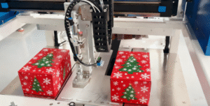 Detailed view of UBL folding box machine (specifically a snap lock bottom box folder): The equipment is processing red Christmas-themed snap lock bottom boxes—adorned with green Christmas tree and white snowflake patterns—with its mechanical components positioned above the finished holiday packages