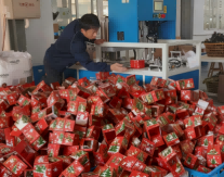 Workers operating a high-speed carton folding machine, forming and stacking Christmas boxes, with a large pile of finished Christmas boxes accumulating.
