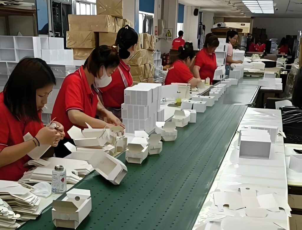 Workers assembling white paper boxes manually on a long production line in a traditional packaging factory, highlighting the contrast with smart factory automation solutions.