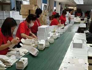Workers assembling white paper boxes manually on a long production line in a traditional packaging factory, highlighting the contrast with smart factory automation solutions.