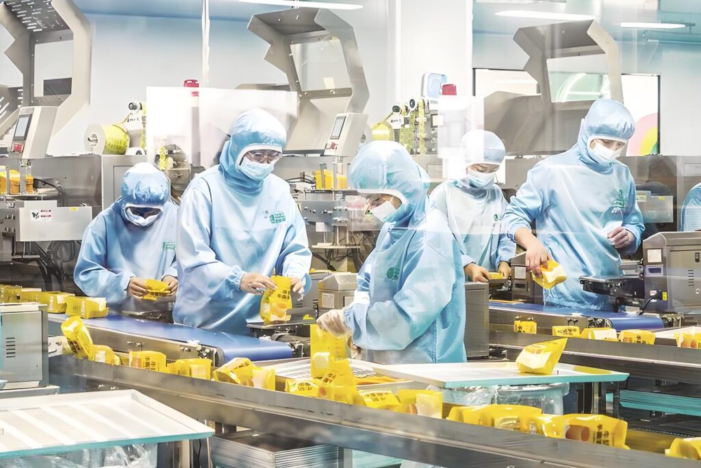 Food factory workers in blue protective gear manually handle yellow-packaged products on a production line, performing manual packaging tasks — this seemingly calm scene embodies the "slow value leak" of manual packaging (e.g., tiny pauses, minor packaging misalignments) highlighted in the article, where unnoticeable small inefficiencies quietly drain resources over time.