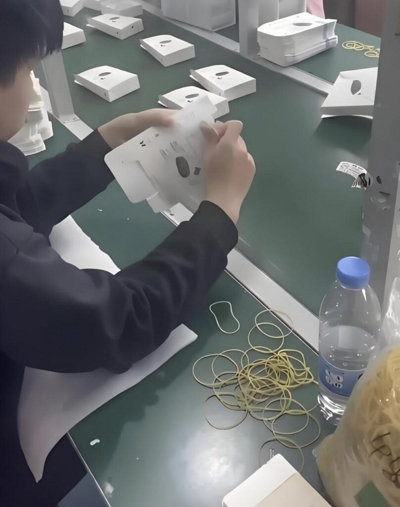 Manual carton folding: worker assembling white tuck-end cartons by hand on a production workbench, highlighting the need for automated carton folding machines
