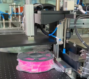 Fully automatic filament vacuum packaging machine in operation, vacuum-sealing a pink 3D printing filament spool in a transparent barrier bag, part of an automated filament packing line for moisture protection.