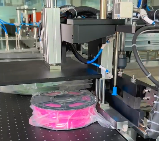 Fully automatic filament vacuum packaging machine in operation, vacuum-sealing a pink 3D printing filament spool in a transparent barrier bag, part of an automated filament packing line for moisture protection.